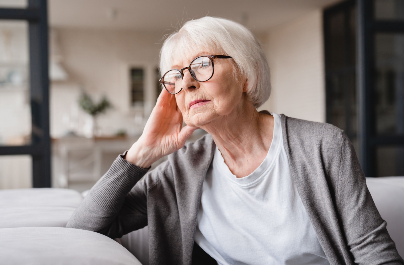 An older adult woman with glasses on sitting on a couch and looking out the window with a serious expression.