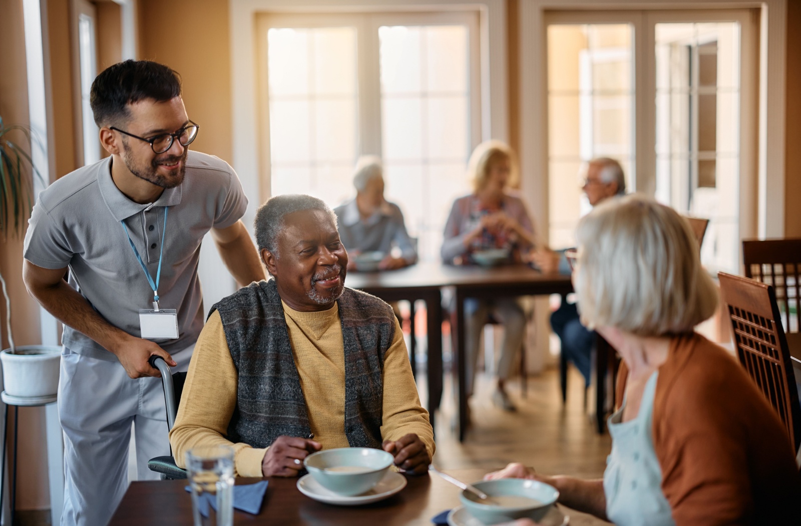 A young male nurse standing behind an older man and older woman sitting at a dining table and they all smile at each other.