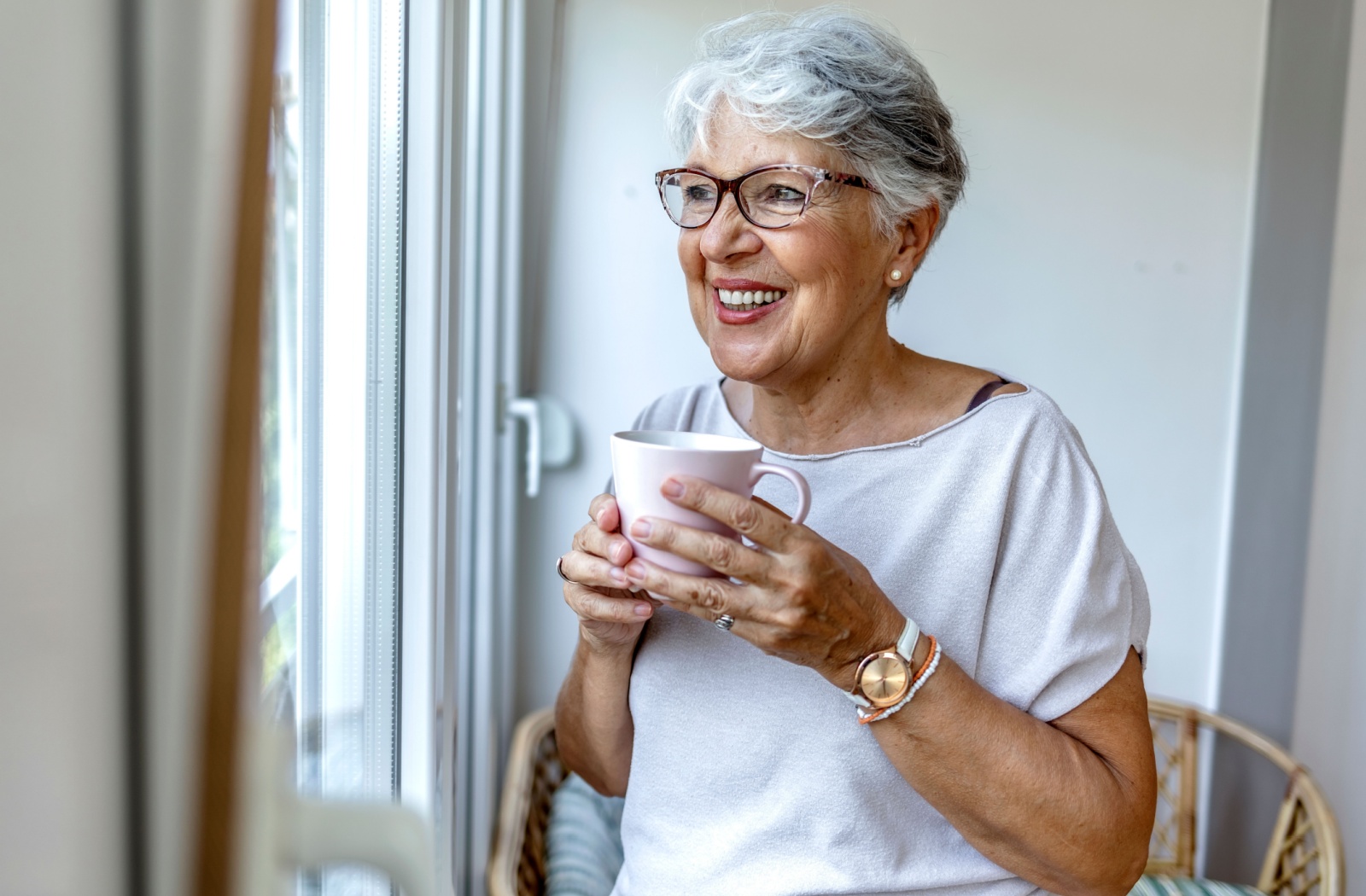 An older woman holding a coffee mug and smiling while looking out of a window.