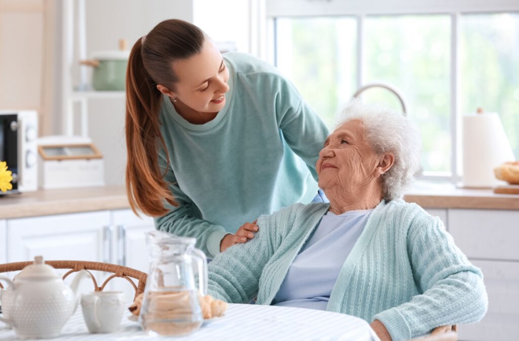 An older woman sitting in a chair looking up at a young, adult woman who is smiling at her.