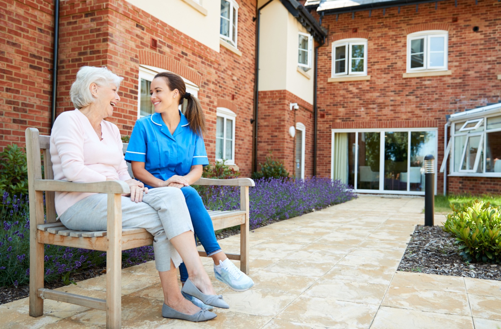 An assisted living caregiver and a resident sit on a bench outside the community.