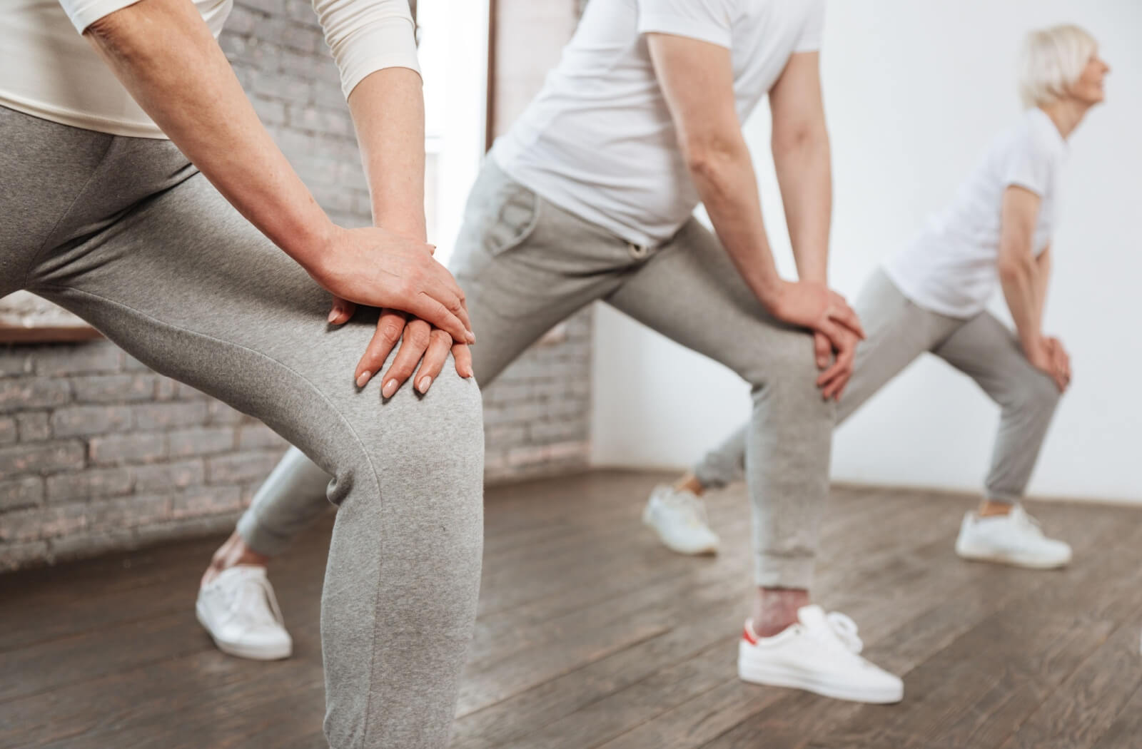 Seniors in workout clothes doing knee strengthening exercises in a bright room with wooden floors.