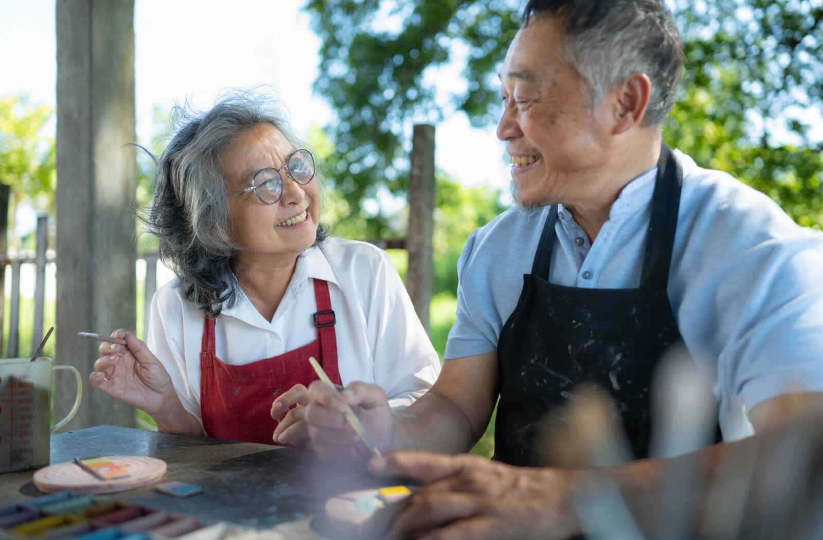 Two older people wearing aprons and holding paint brushes painting together at an outdoor table.