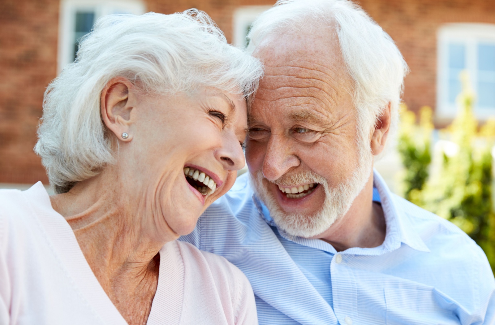 An older couple touches their foreheads together while laughing outside in the courtyard of their assisted living community
