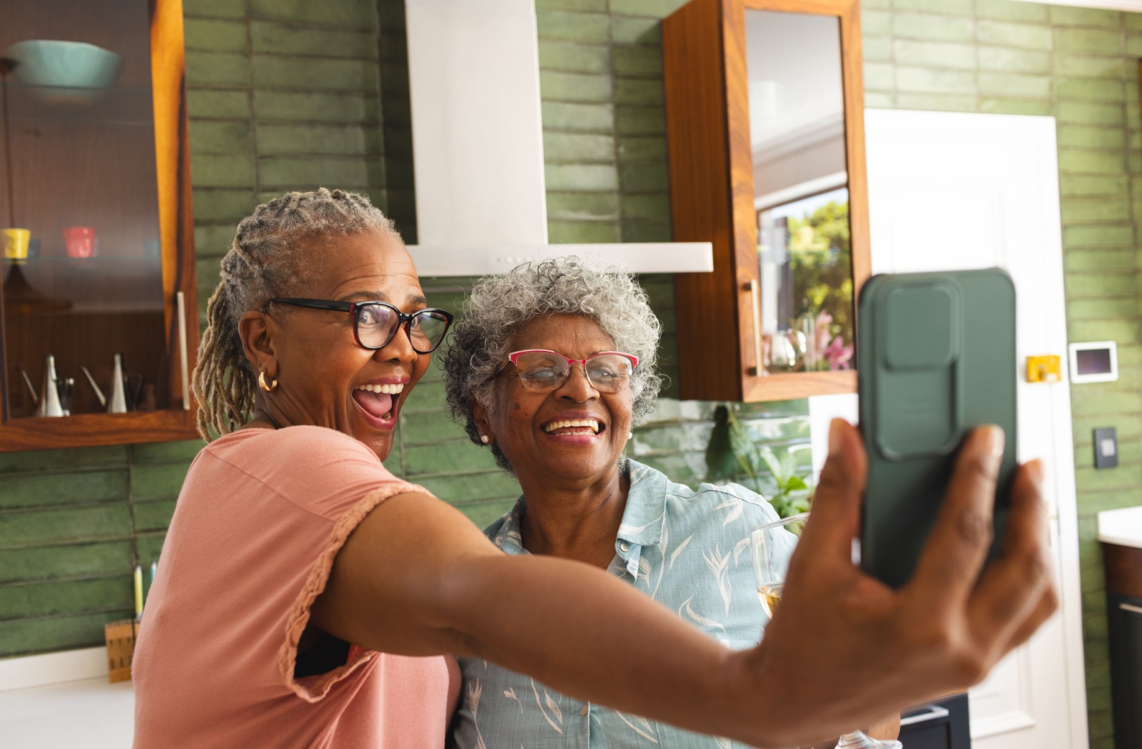 Two seniors smile widely as they take a picture together in a cool-colored common space of their assisted living home